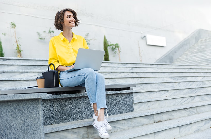 A woman sitting on a set of steps smiling while typing on a laptop 