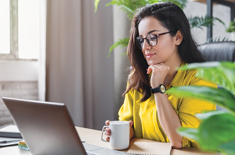 A woman sitting at a desk looking at her laptop while holding a coffee cup