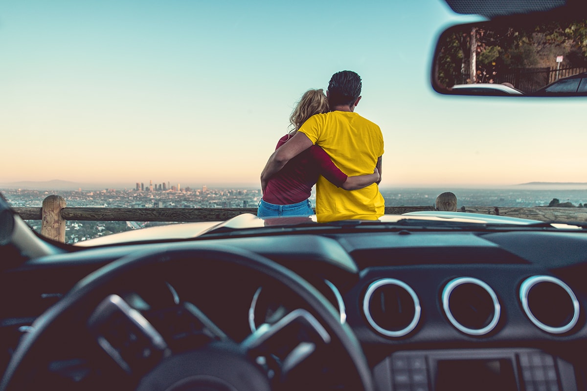 A couple hugging while sitting on hood of a car and looking out over city skyline