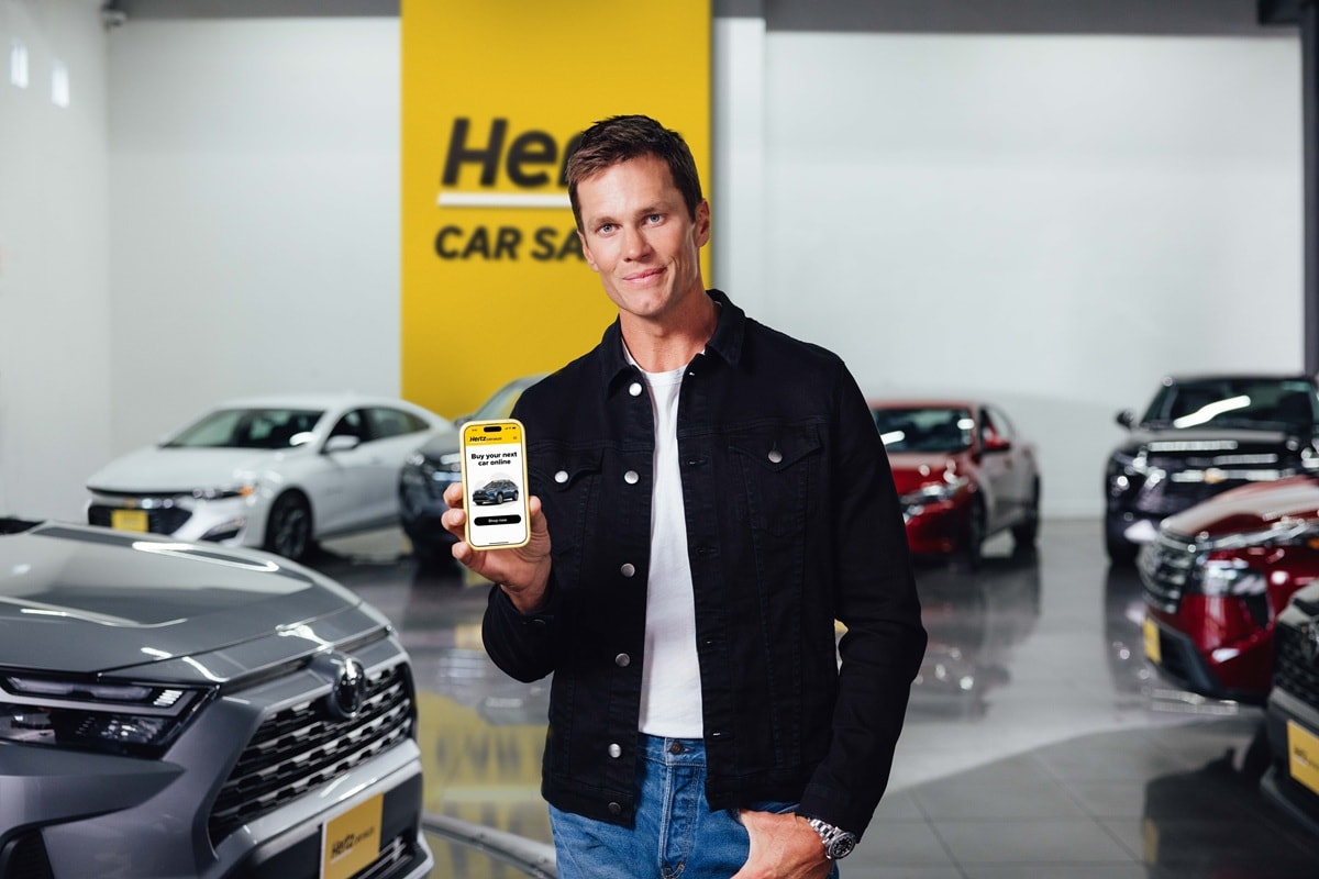 Tom Brady holding out a phone surrounded by Hertz Car Sales vehicles