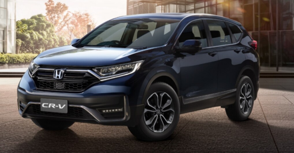 A deep blue 2023 Honda CR-V sits parked on concrete in front of a glass building with the sun shining and some trees in the background