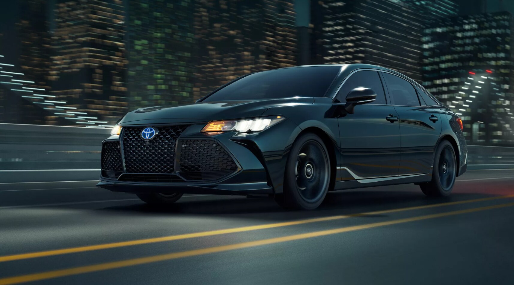 A black 2022 Toyota Avalon Hybrid speeds past the viewer on a dusky night with a cityscape in the background