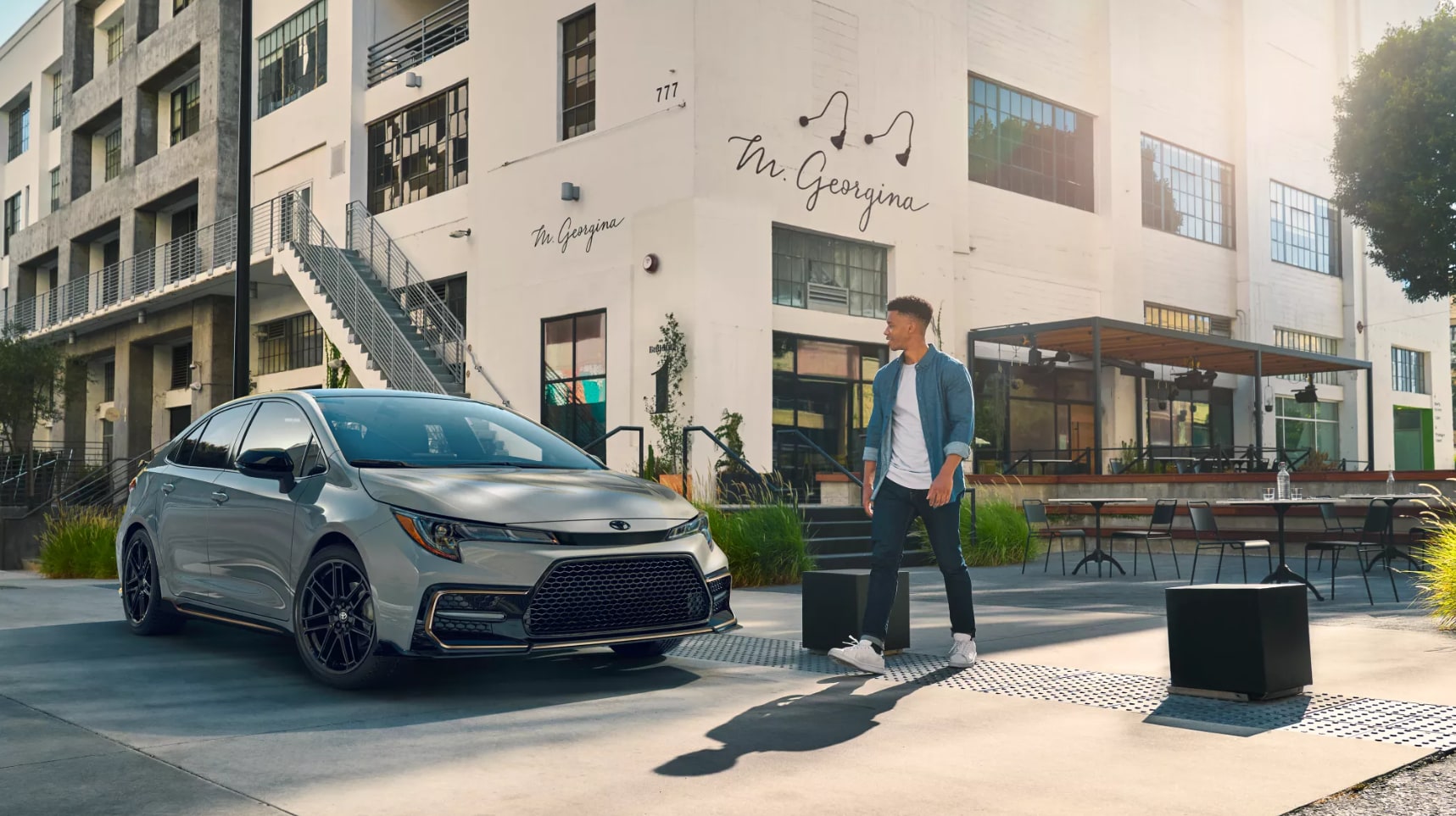a man walks in front of a parked light gray 2022 Toyota Corolla Hybrid as it sits parked in front of an urban warehouse/restaurant