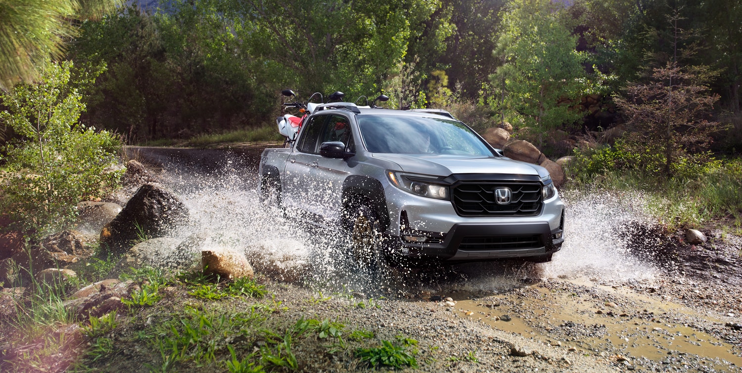 silver Honda Ridgeline tuck splashing in a wooded area with an ATV in the bed