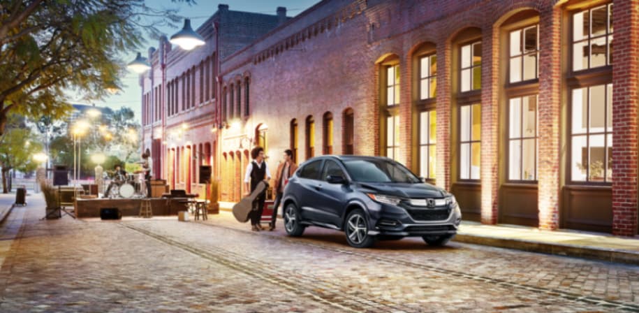 black Honda HR-V SUV parked on a cobblestone street in front of a brick building, musicians approaching