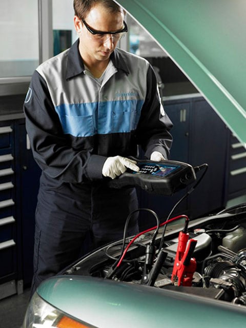 Honda Battery Replacement at Honda of Salem A Honda service technician standing over a vehicle engine bay testing a car battery.
