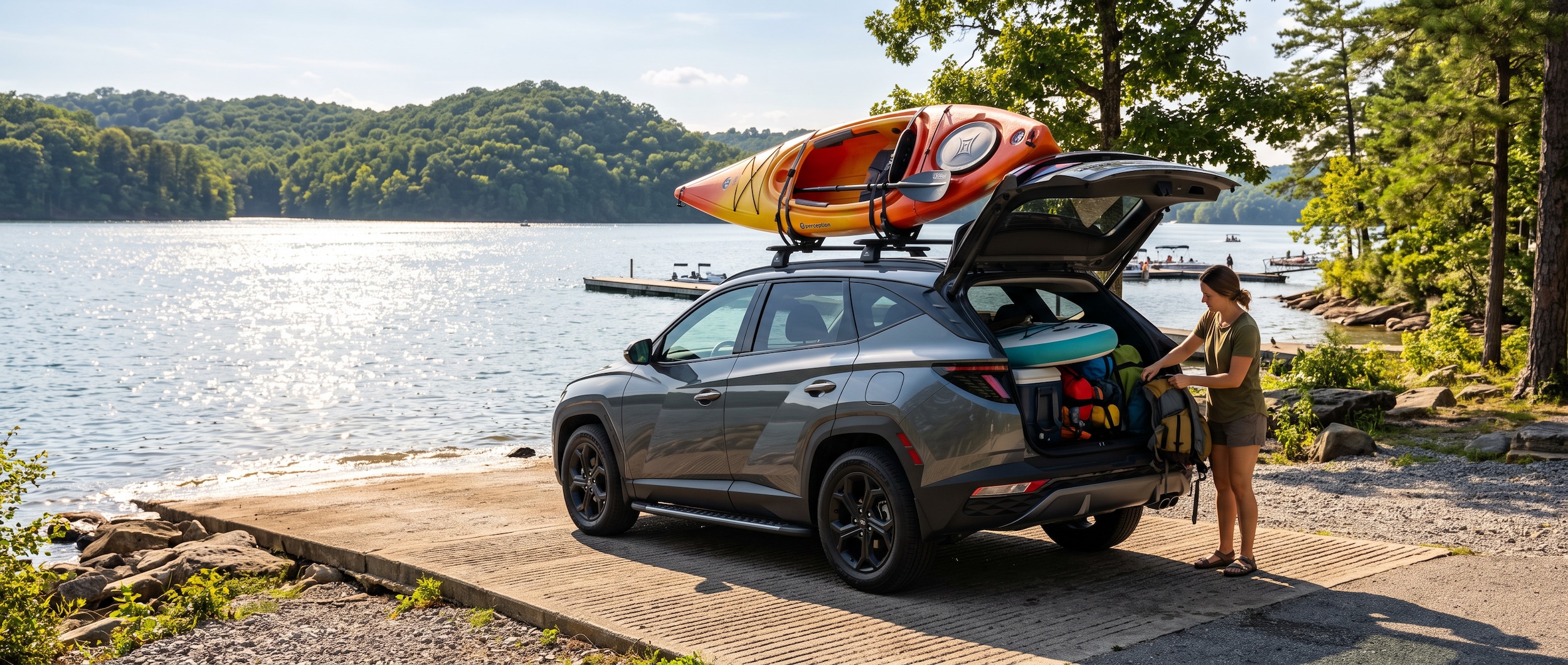 A 2026 Hyundai Tucson XRT in a deep green or gray finish parked at a Tennessee lakeside boat ramp with sun on the water behind it, late-summer light, gear loaded in the cargo area, and a kayak on the roof