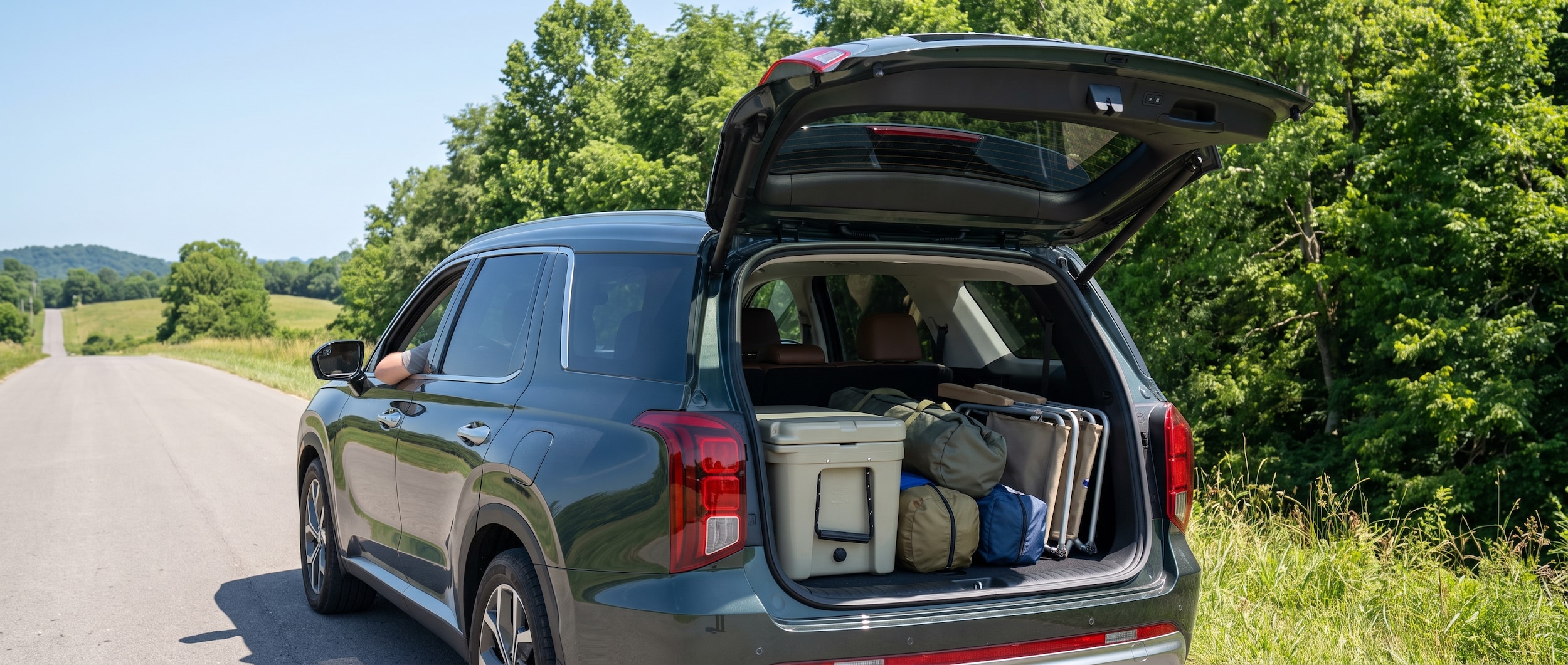 A 2026 Hyundai Palisade with rear liftgate open, loaded with a cooler, soft duffel bags, and folded camping chairs, parked on a sun-drenched Middle Tennessee road with green summer trees in the background