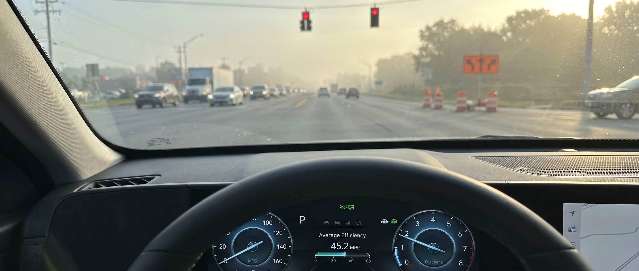 A 2026 Hyundai Kona in a modern exterior color sitting at a red light on a wide, busy Nashville-area arterial road during a hazy summer morning, construction signage visible in the background, dashboard in sharp focus showing an efficiency readout