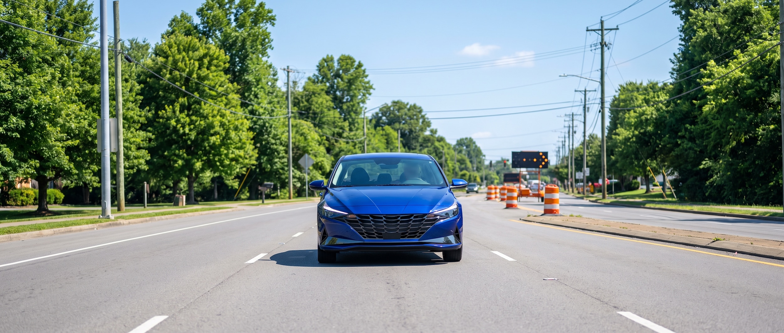 A 2026 Hyundai Elantra in silver or blue driving on a wide four-lane surface road in suburban South Nashville, Tennessee in summer morning light, with construction signage visible in the background and lush green trees lining the roadway