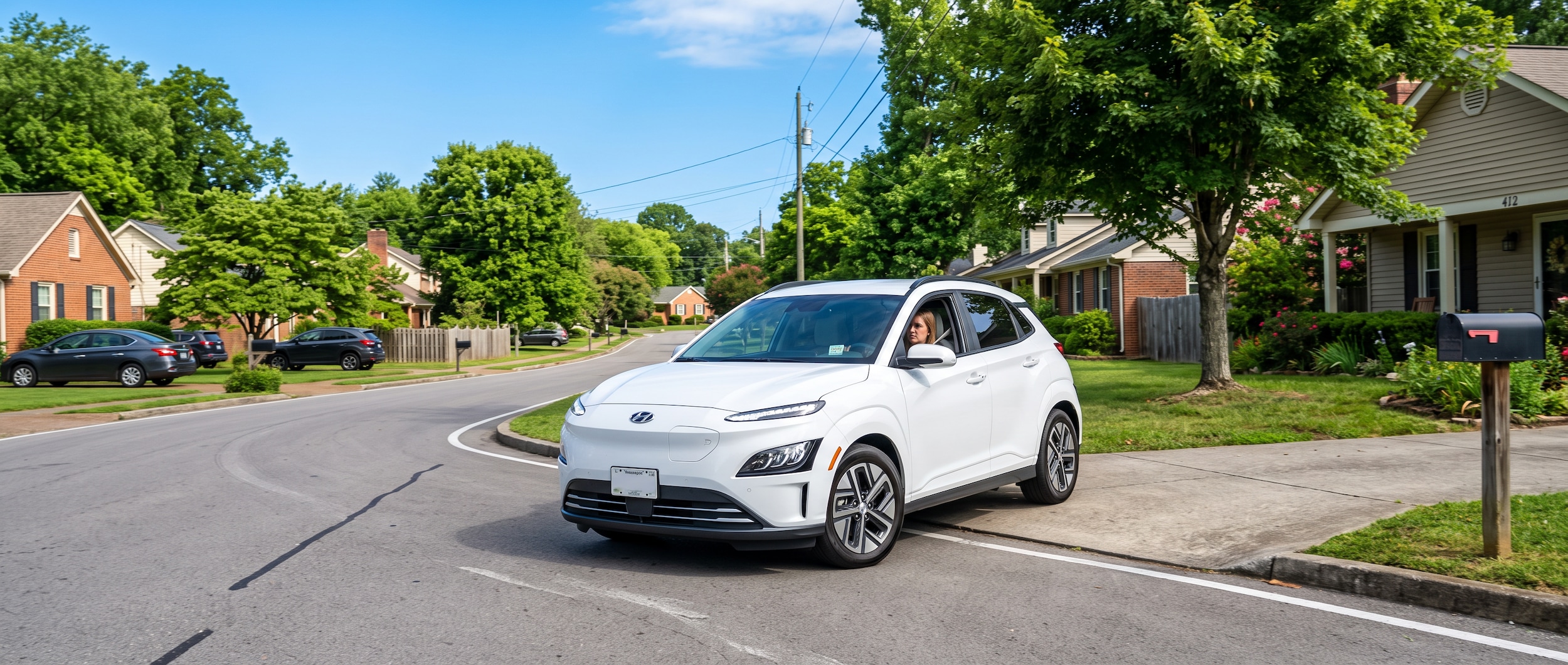 A 2025 Hyundai Kona Electric in a deep blue or Atlas White finish pulling out of a suburban South Nashville neighborhood on a summer morning, residential streets visible in the background, bright Tennessee sky overhead