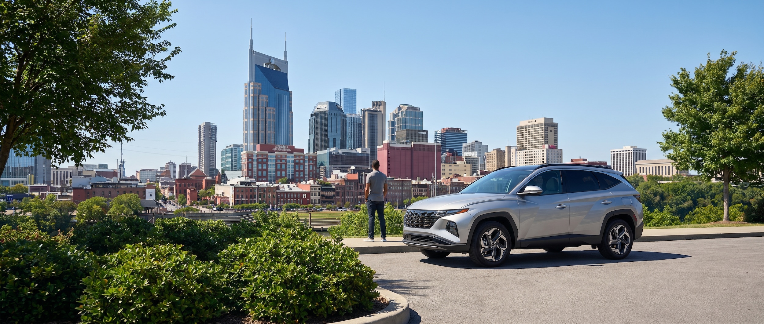 A silver Hyundai Tucson Hybrid parked near a scenic overlook with the Nashville skyline visible in the background on a clear sunny day, conveying an urban lifestyle feel with natural green surroundings.