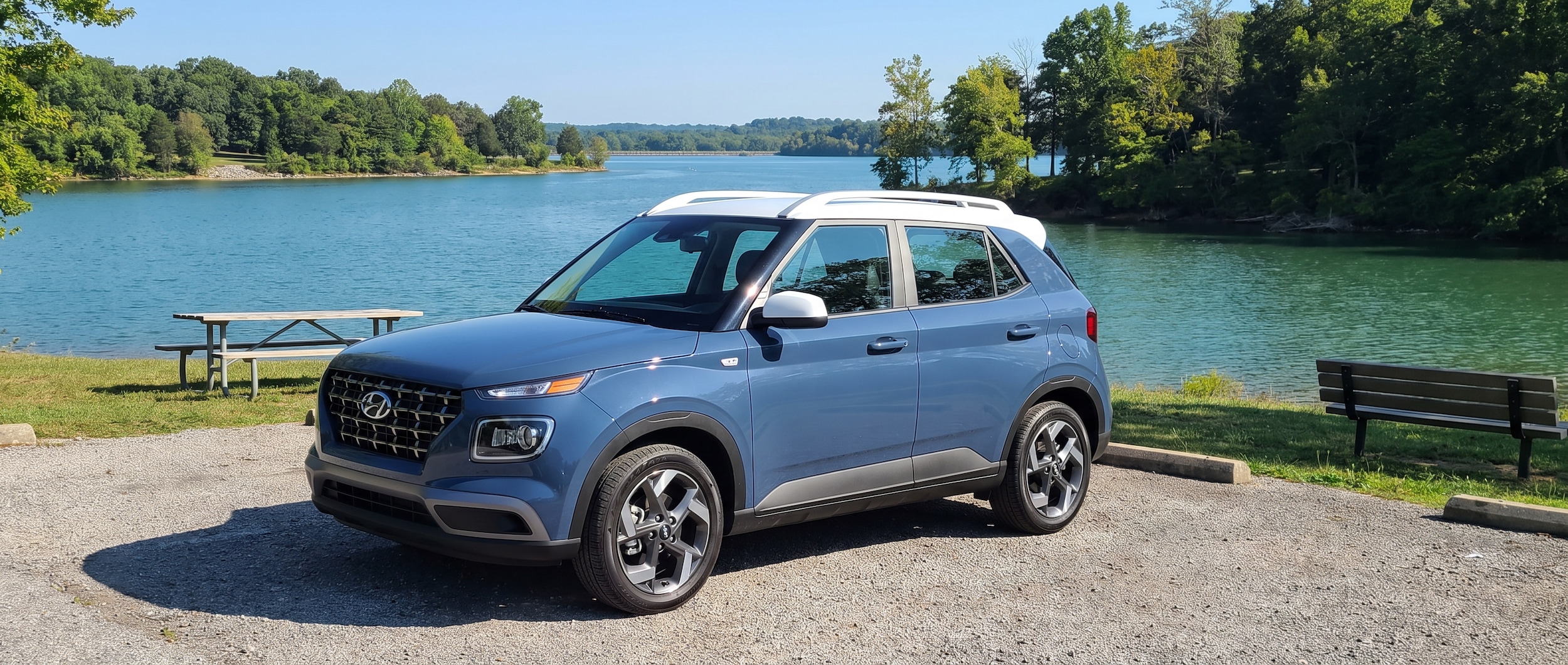 A 2026 Hyundai Venue SEL in Denim blue with contrasting white roof parked at a lakeside day-use area at Long Hunter State Park on a bright August morning, calm blue-green water and Tennessee tree line visible behind the vehicle