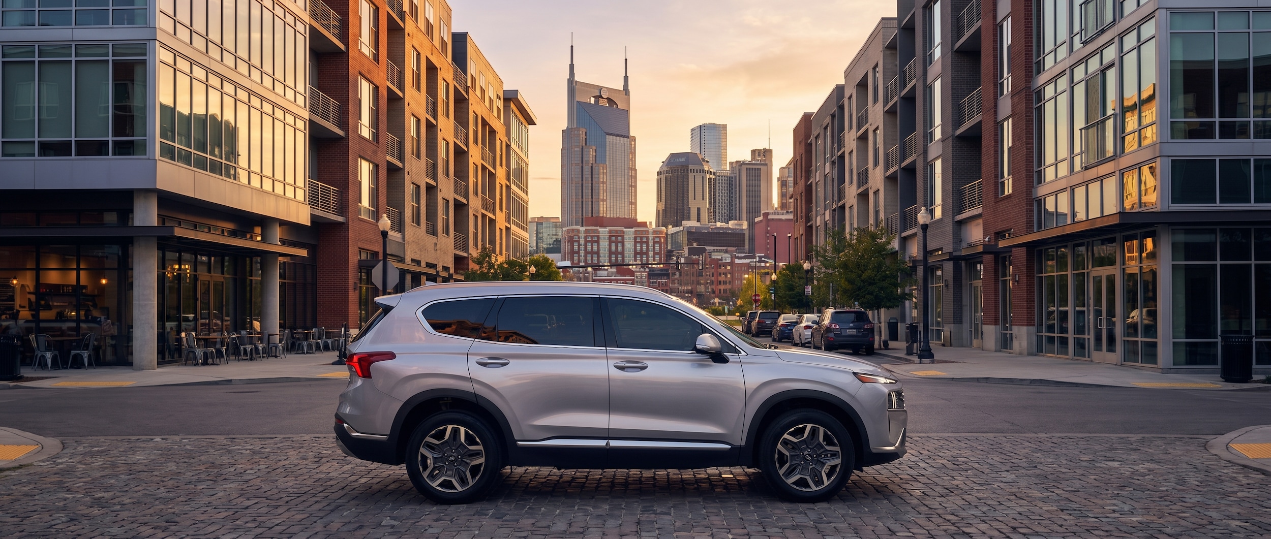 A silver Hyundai Santa Fe Hybrid parked in an urban Nashville setting near modern mixed-use buildings with city skyline visible in the background during golden hour