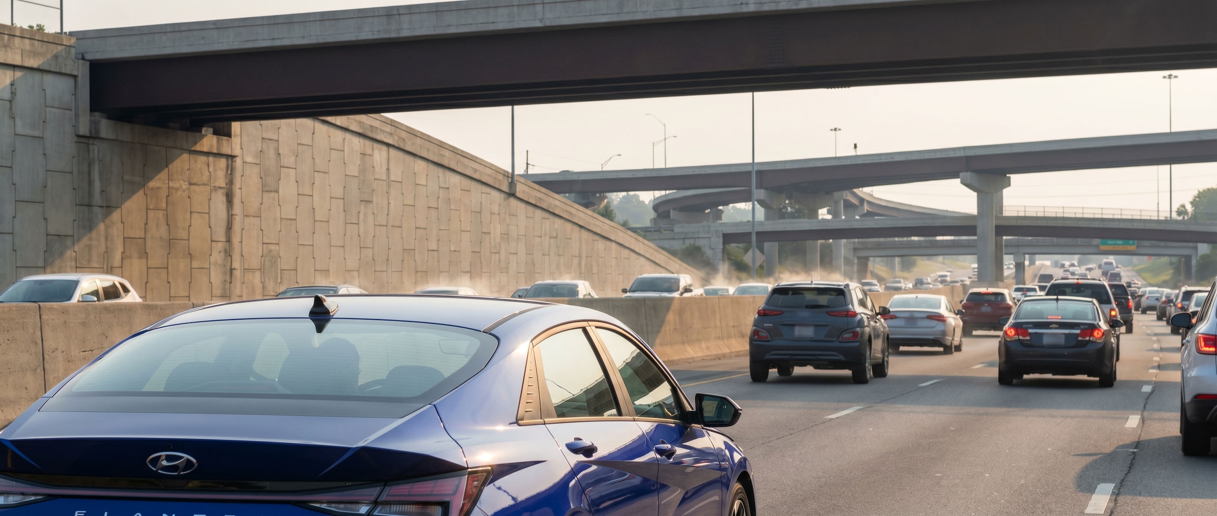 A 2026 Hyundai Elantra Hybrid in a modern color (white or Intense Blue) on a Nashville-area highway during a summer morning commute, with a hazy blue sky and urban highway infrastructure visible in the background, conveying heat and congestion