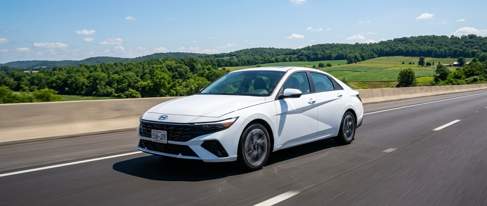 A 2026 Hyundai Elantra in silver or white rolling along a clear interstate highway in rural Tennessee or Kentucky in summer daylight, green rolling hills and blue sky visible, shot from a low three-quarter front angle showing the sedan's athletic profile