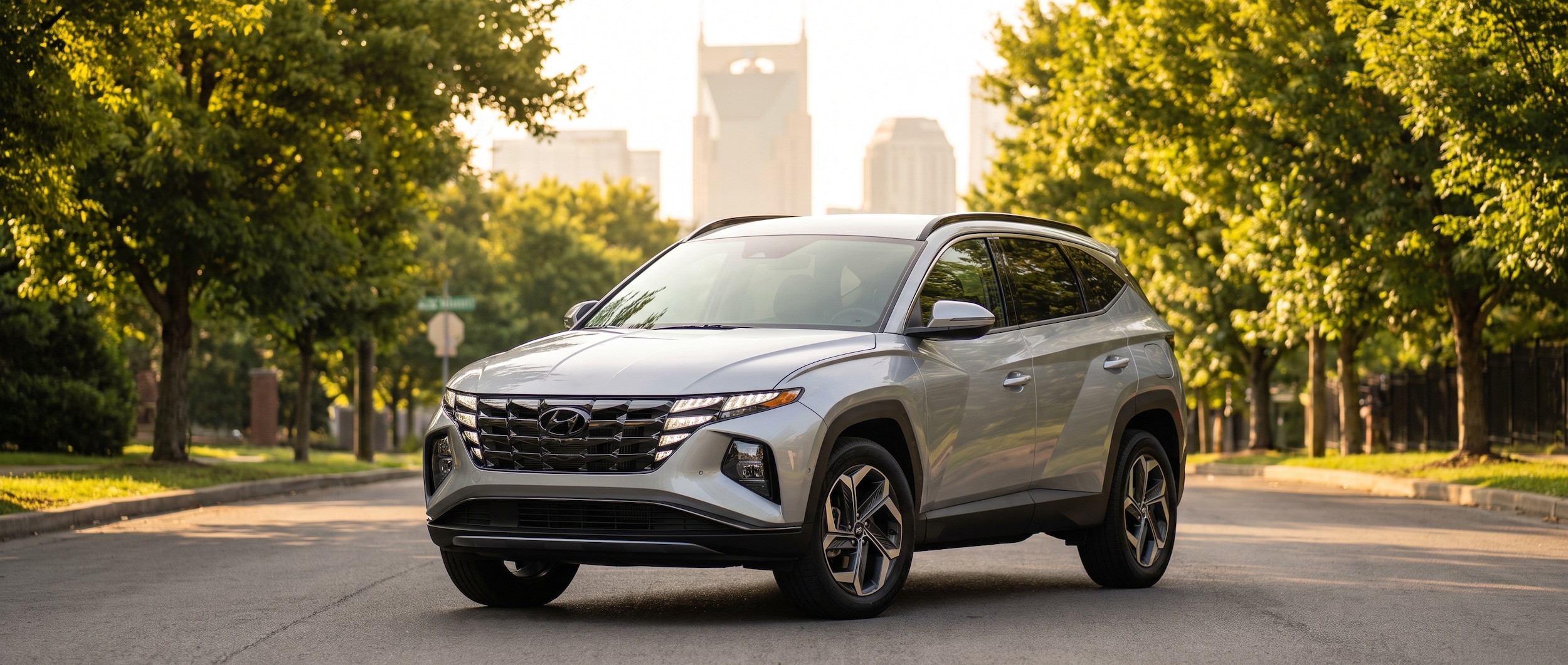 A 2026 Hyundai Tucson Hybrid in silver or Shimmering Silver Pearl parked on a tree-lined Nashville street in mid-July morning light, with a blurred cityscape of downtown Nashville visible in the background, front three-quarter view showing the refreshed front fascia and 18-inch alloy wheels