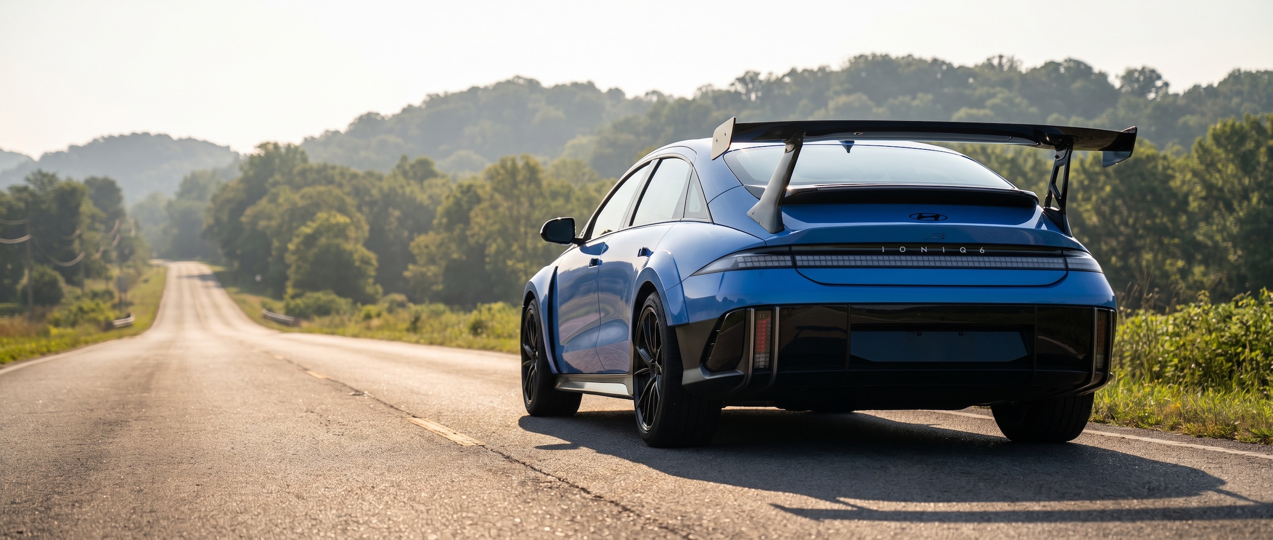 A 2026 Hyundai IONIQ 6 N in Performance Blue Pearl parked on a stretch of sunlit Tennessee highway, low-angle shot emphasizing the swan-neck rear wing and wide rear fenders, summer haze visible in the background