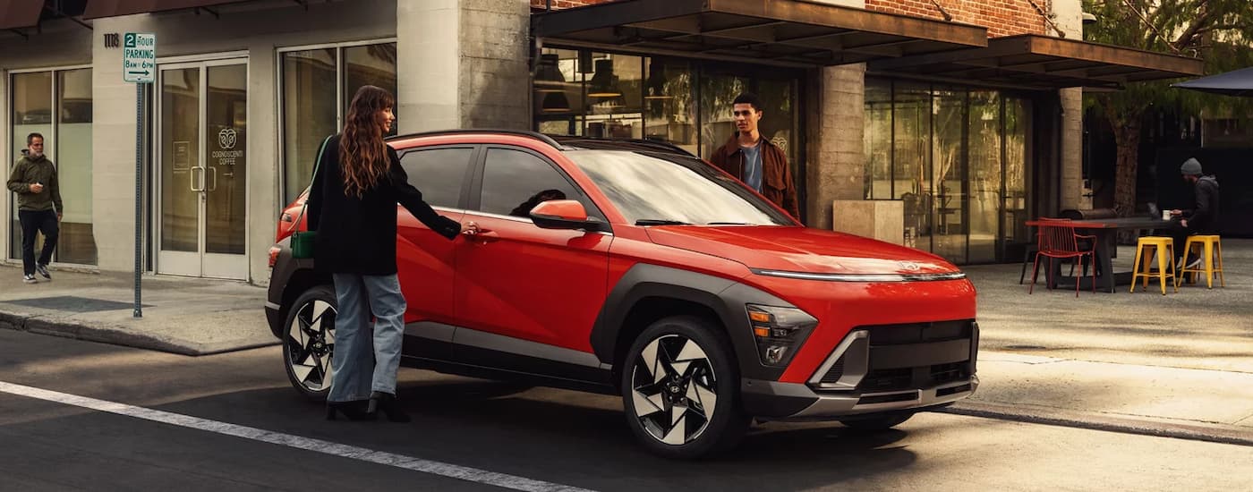 Two people walking by a red 2025 Hyundai Kona for sale near Weddington parked by a curb.