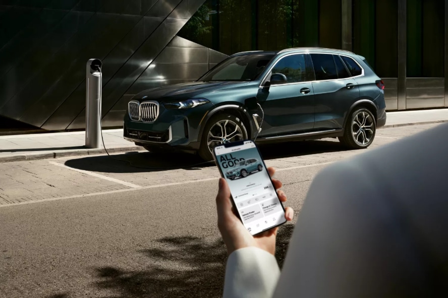 A teal BMW X5 charging at the curb while a person checks their My BMW app
