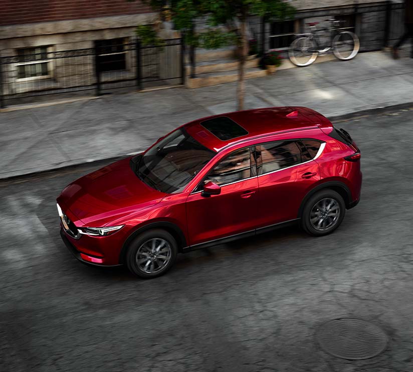 Model Features of the 2021 Mazda CX-5 at Jack Giambalvo Mazda | Aerial Shot of Red 2021 Mazda CX-5 Driving Through City Past Townhomes