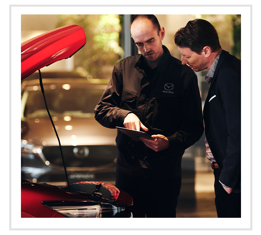 Everything You Need to Know about Your Car Battery at Jack Giambalvo Mazda | Mazda Service advisor showing customer information about car battery while the hood of a red Mazda is open   