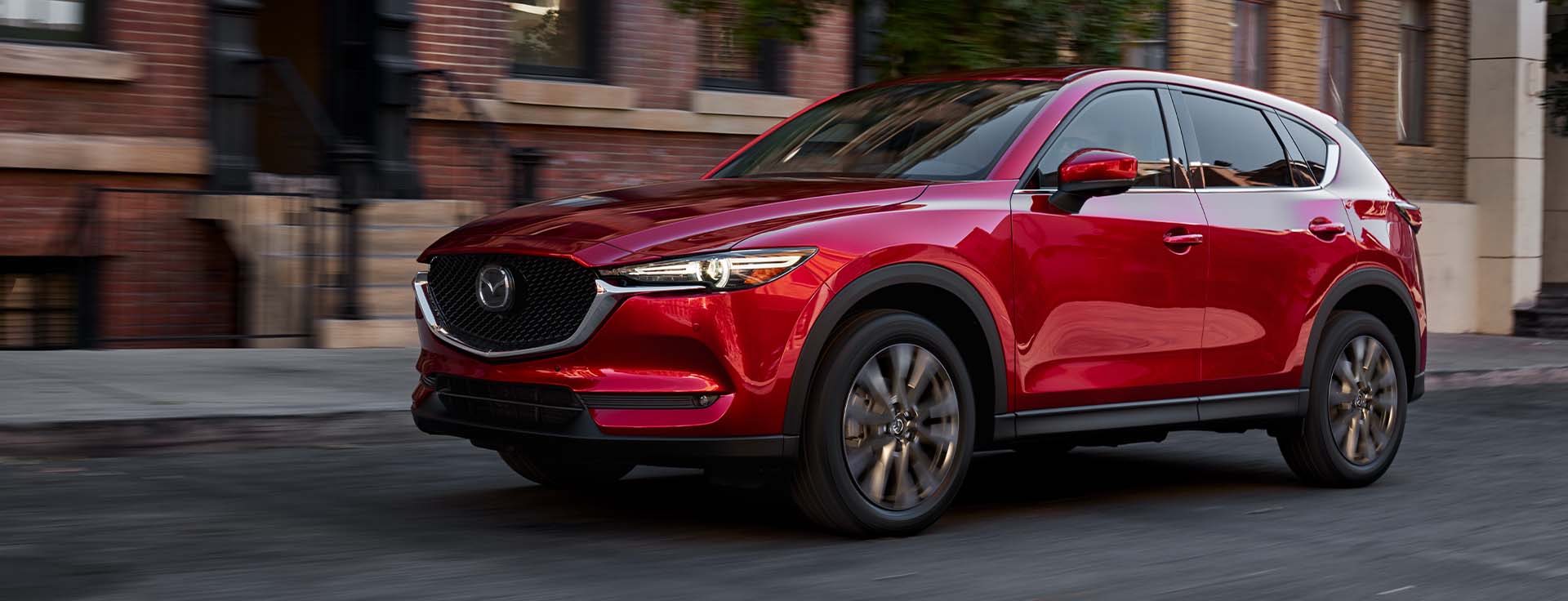 Model Features of the 2021 Mazda CX-5 at Jack Giambalvo Mazda | Red 2021 Mazda CX-5 Driving Past Townhomes in City
