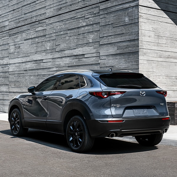 The 2022 Mazda CX-5 vs. The 2022 Mazda CX-30 at Jack Giambalvo Mazda | Rear View of Dark Silver 2022 Mazda CX-30 Parked Next to Stone Building