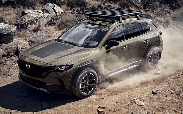 Model Features of the All-New 2023 Mazda CX-50 at Jack Giambalvo Mazda | Zircon Sand 2023 Mazda CX-50 Facing Left Angled Towards Camera Driving on Dirt Road with Large Boulders