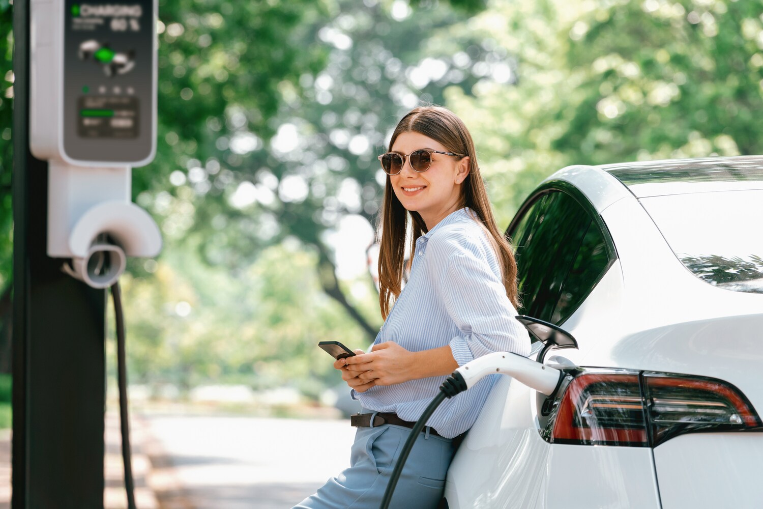 Woman standing against electric vehicle on her phone while waiting for her EV to charge at a public charging station near Roswell, GA
