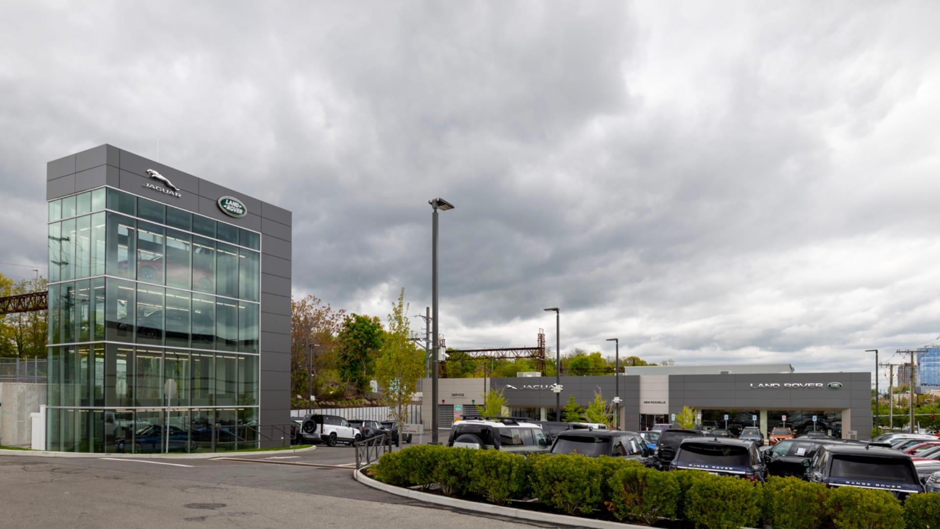 Grey Land Rover New Rochelle dealership building with a cloudy sky in the background. Vehicles can be seen through the large glass windows in a multi story display structure on the left, and the dealership building is visible on the right with many vehicles in the lot surrounding it.