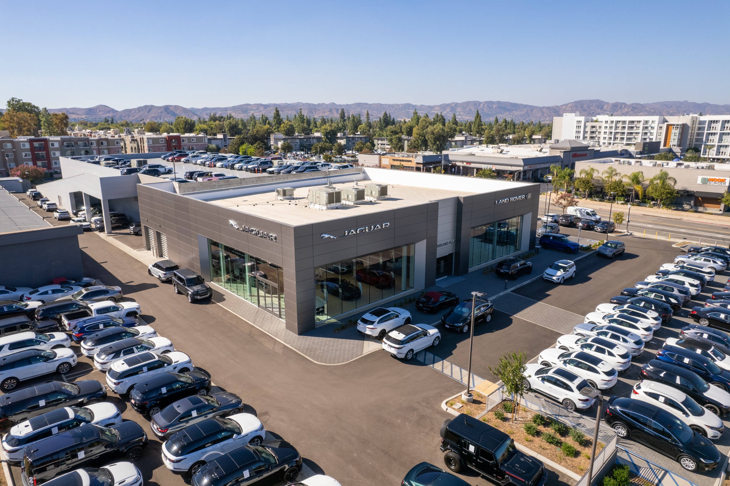 Aerial view of Jaguar Woodland Hills car dealership surrounded by vehicles in the parking lot. Evergreen trees and are in the background, with mountains further in the background.