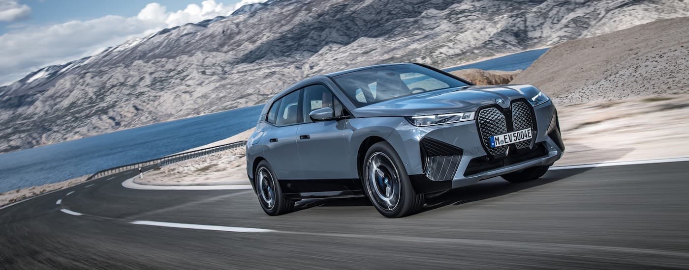A newer BMW EV, a 2022 BMW iX xDrive50, is shown rounding a corner with a lake and mountains in the background.