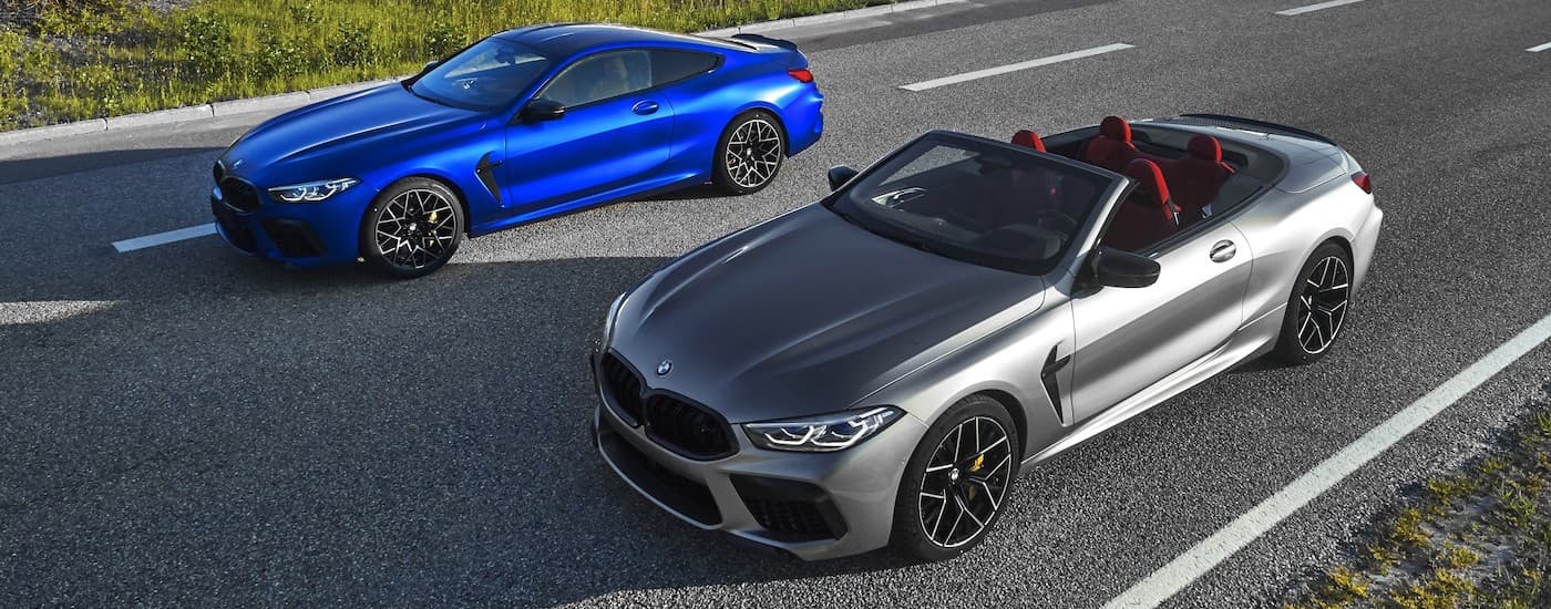 A blue 2020 BMW M8 coupe and a silver convertible are parked next to each other on an empty road.