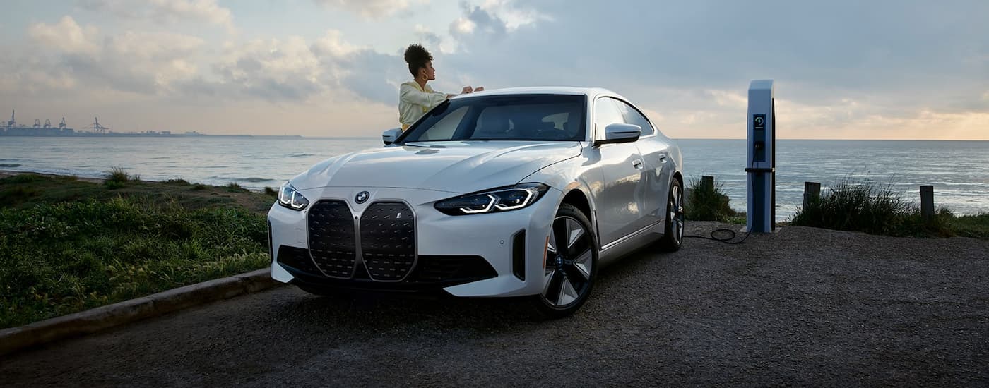 A woman is shown standing next to a white 2023 BMW i4 Gran Coupe near the ocean.