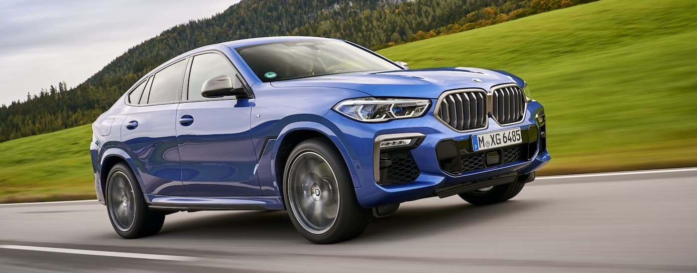 A blue 2021 BMW X6 M50i is shown driving past a field.