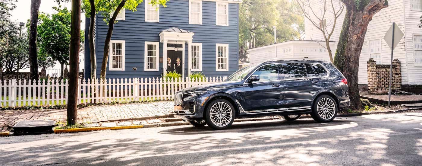A dark blue 2021 BMW X7 is shown parked in front of a house and white picket fence.