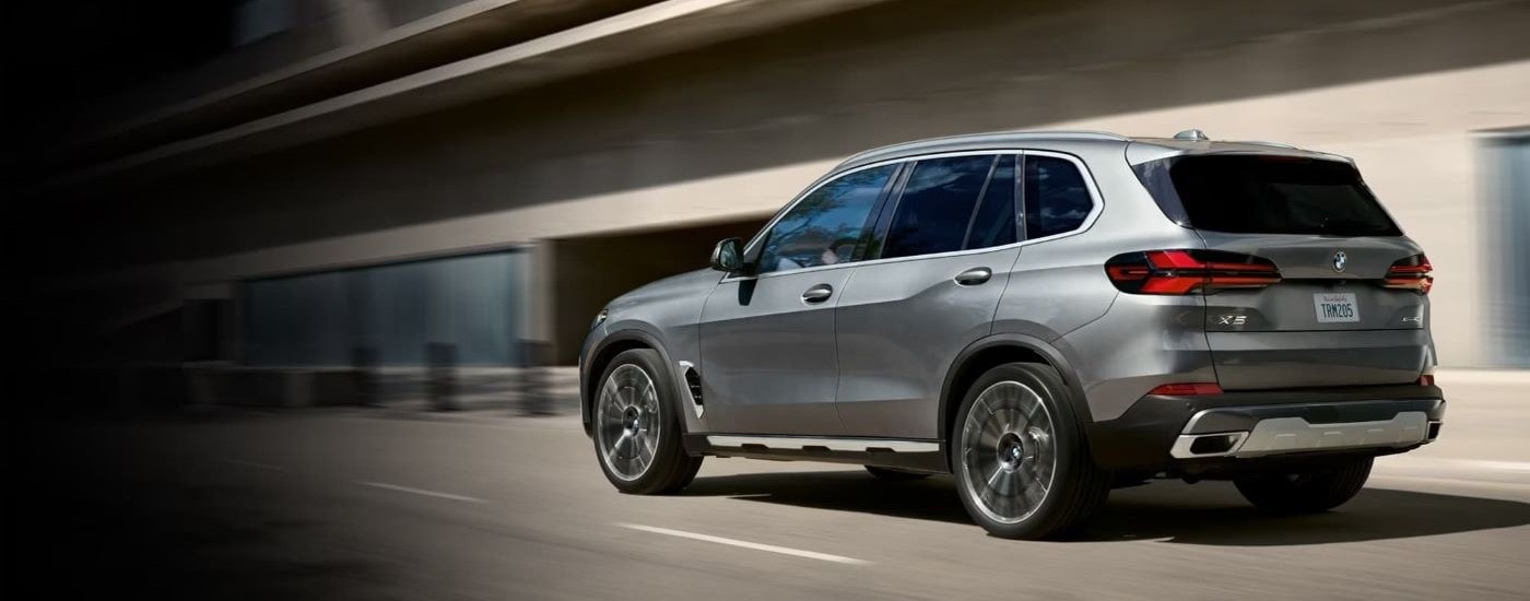 A silver 2024 BMW X5 is shown from the side in front of a building.