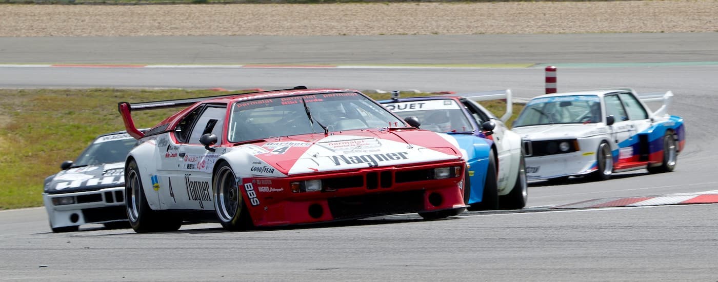 Multiple older BMW M Series vehicles are driving on a track during an anniversary race.