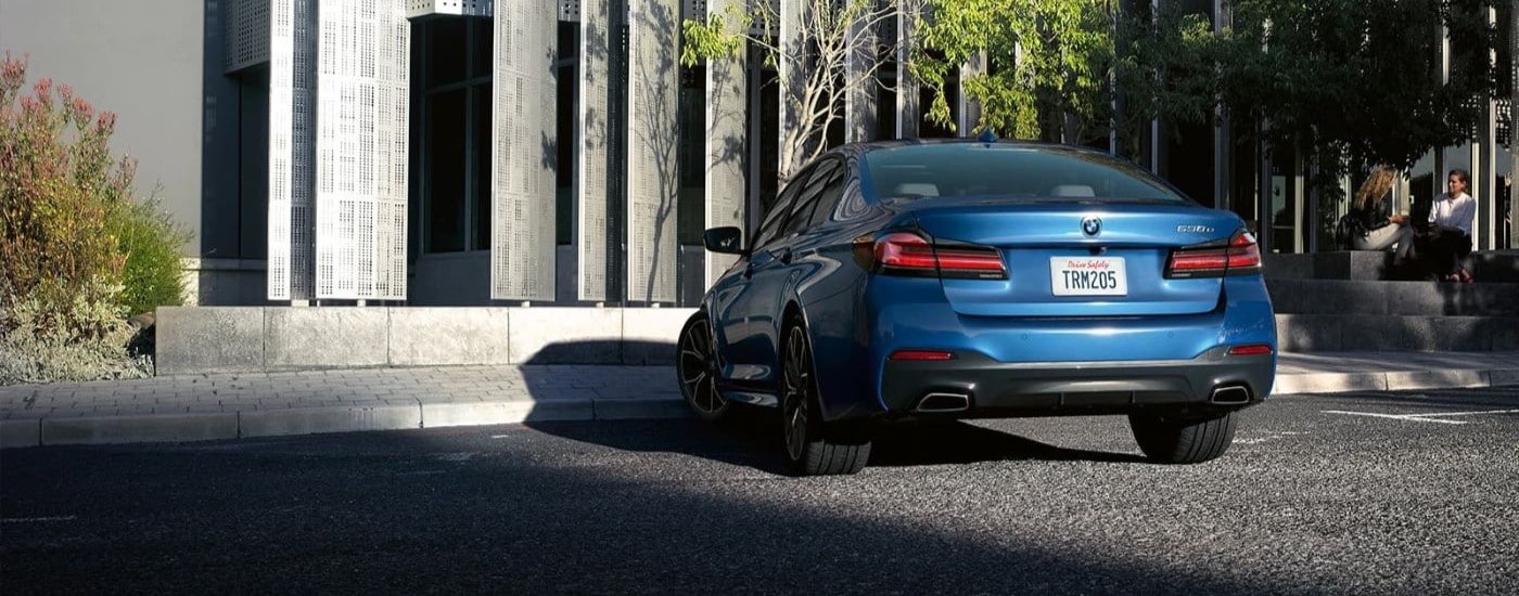 A blue 2023 BMW 5 Series is shown from the rear parked in front of a modern building.