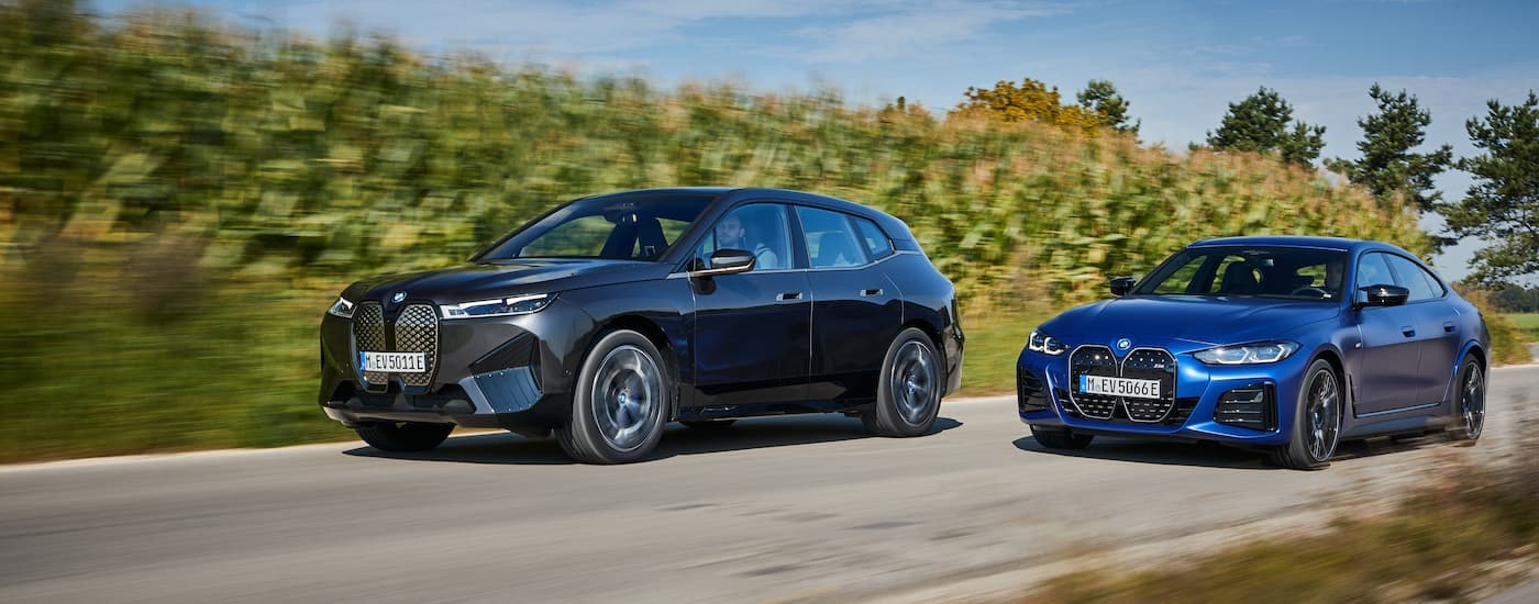 A dark grey 2022 BMW iX and a blue i4 are shown driving past a corn field.