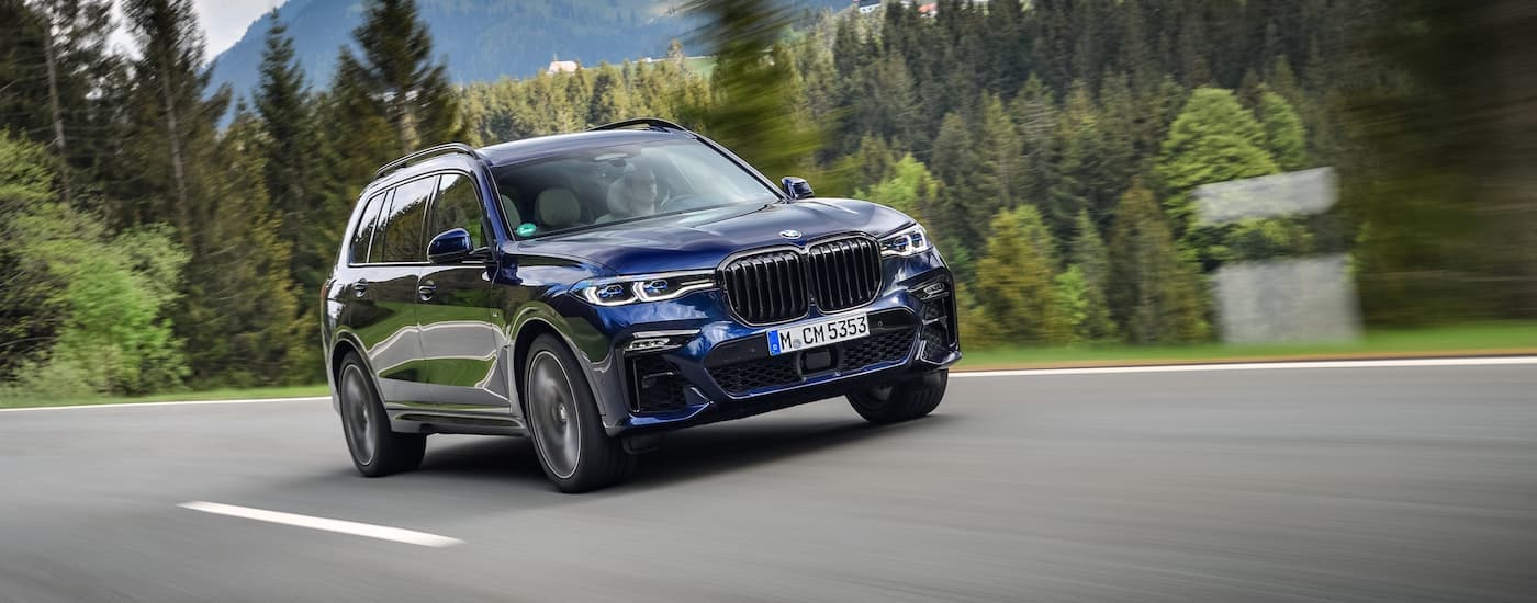 A blue 2022 BMW X7 m50i is shown driving on a tree-lined road after visiting a Dayton BMW delaer.