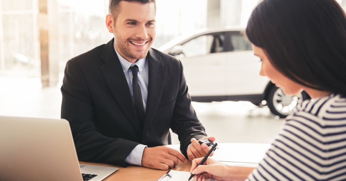 Customer signing vehicle purchase paperwork while salesperson hands over car keys in a dealership showroom.