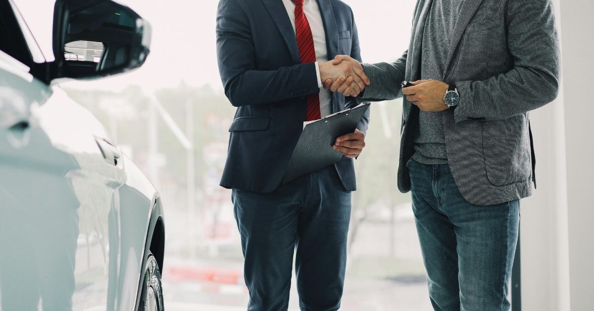 Car salesperson and customer shaking hands beside a vehicle in a bright dealership showroom.