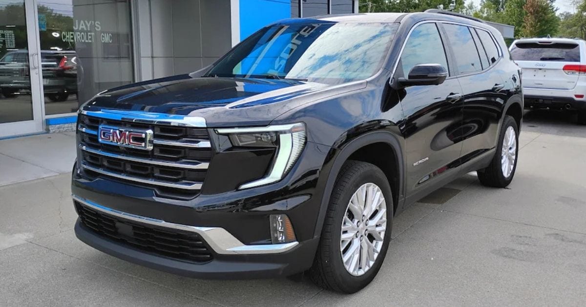 Front three-quarter view of a black 2024 GMC Acadia Elevation SUV parked on a dealership lot outside Jay’s Chevrolet GMC, with a white SUV in the background.