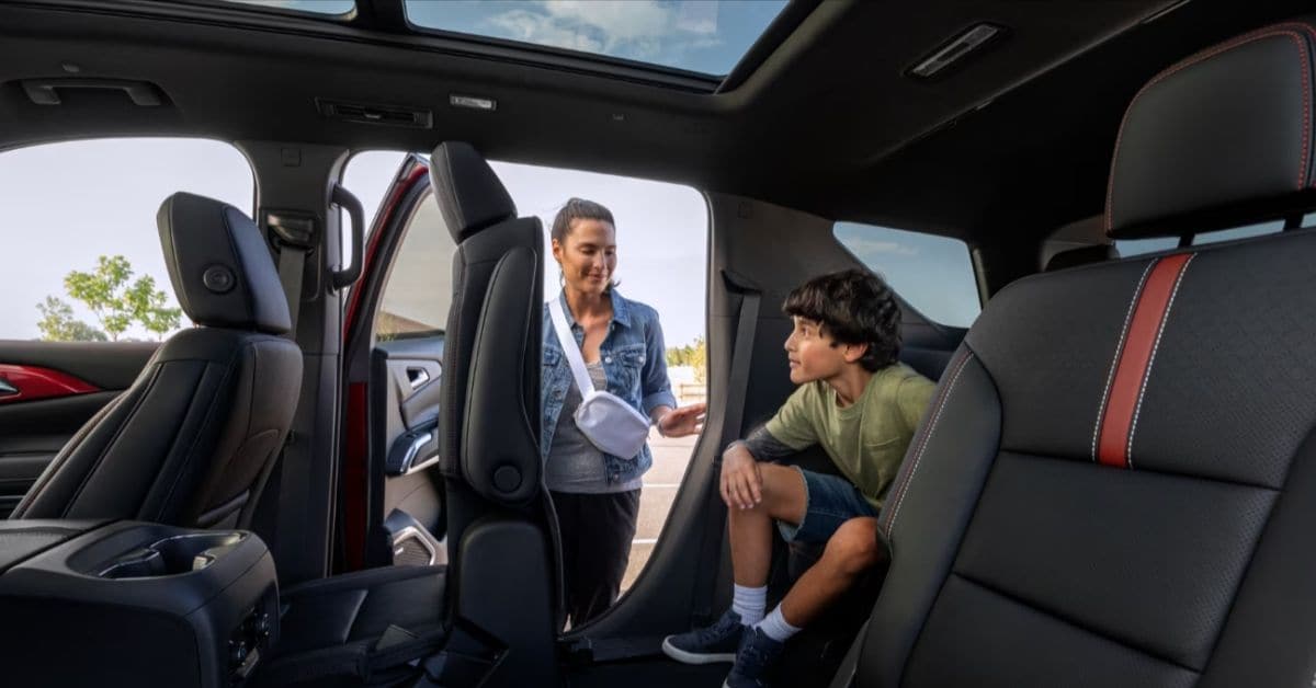A mother smiles at her young son seated in the third row of a spacious 2027 Chevrolet Traverse SUV with black leather seats, red stitching, a folded second-row seat for easy access, and a panoramic sunroof overhead.