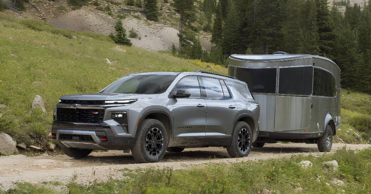 A gray 2027 Chevrolet Traverse Z71 SUV tows a silver travel trailer on a dirt road in a mountainous forest setting.