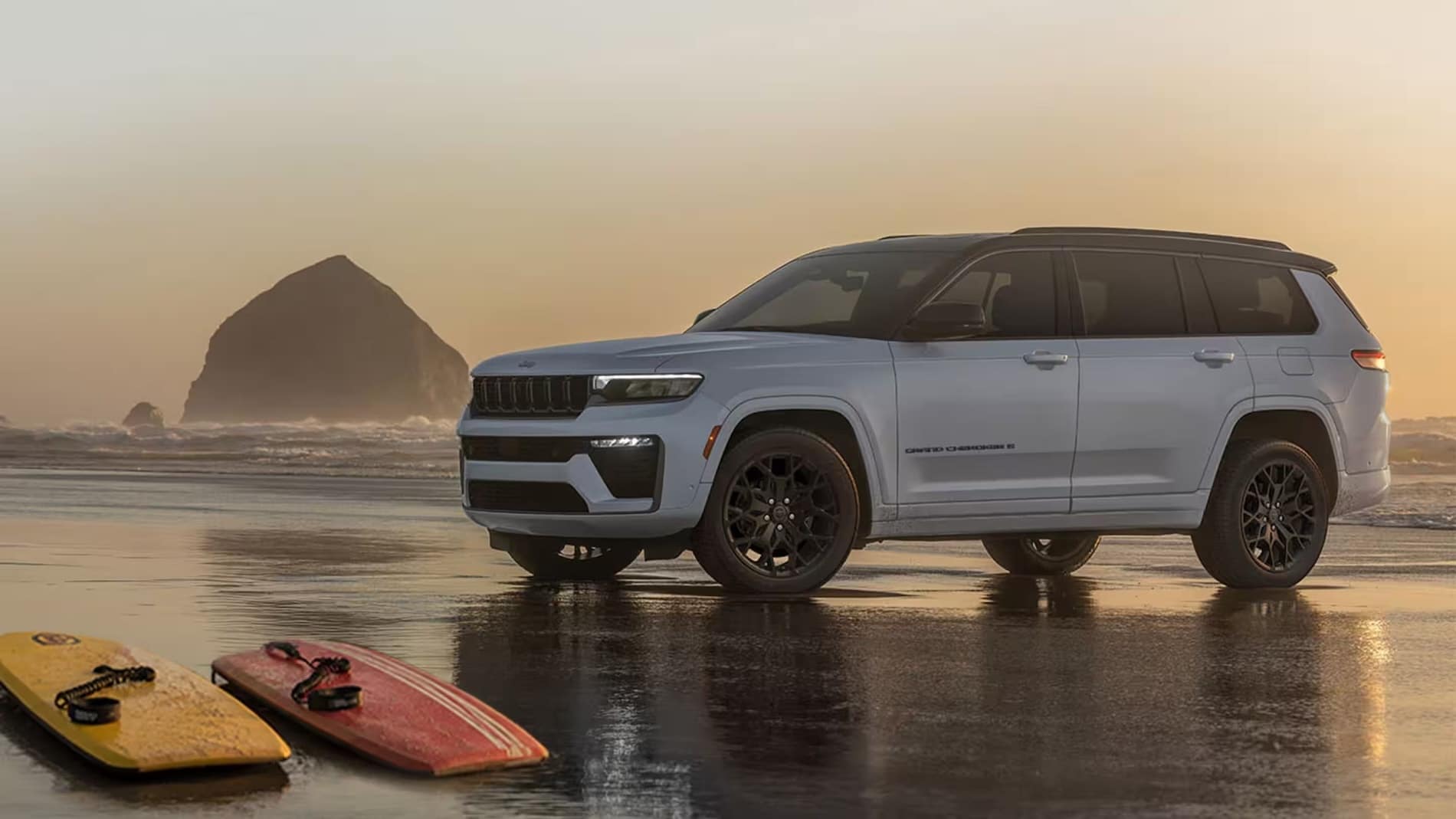 White 2027 Jeep Grand Cherokee parked on a beach at sunset, side profile, with surfboards in the foreground