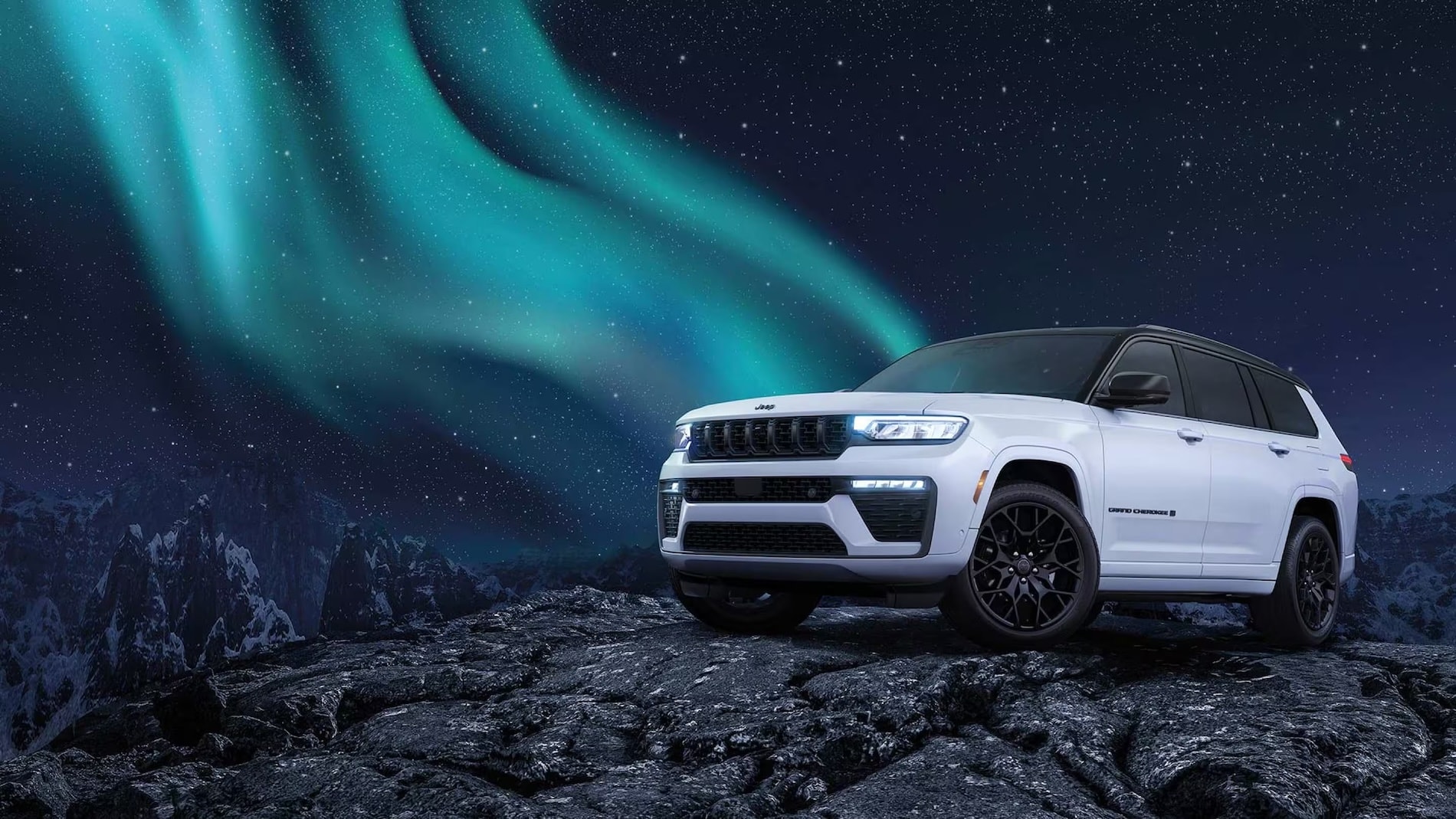 White 2027 Jeep Grand Cherokee front three-quarter view on a rocky overlook under the northern lights