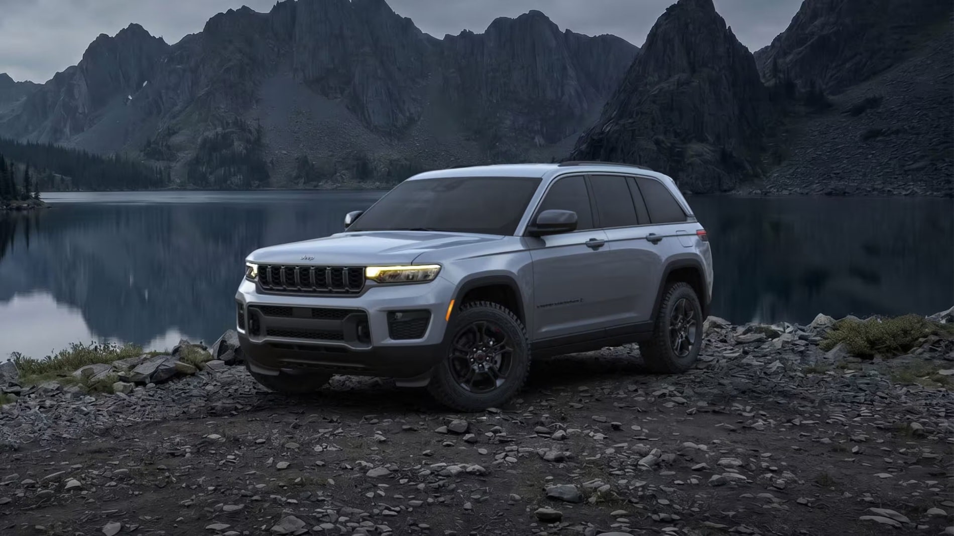 Silver 2027 Jeep Grand Cherokee front three-quarter view beside a mountain lake at dusk